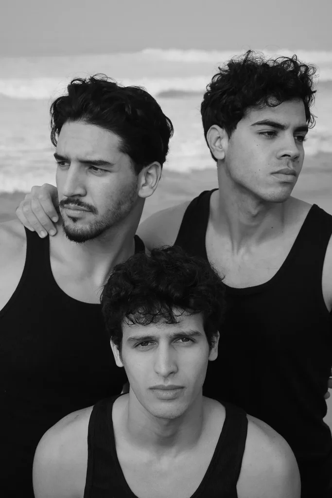 Portrait of three young men in black tank tops on the beach – black and white photograph by Davide Edoardo