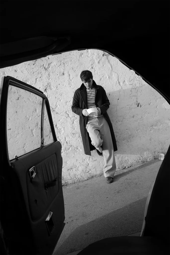 Young man leaning against a wall, photographed through the open door of a vintage car – black and white photo by Davide Edoardo