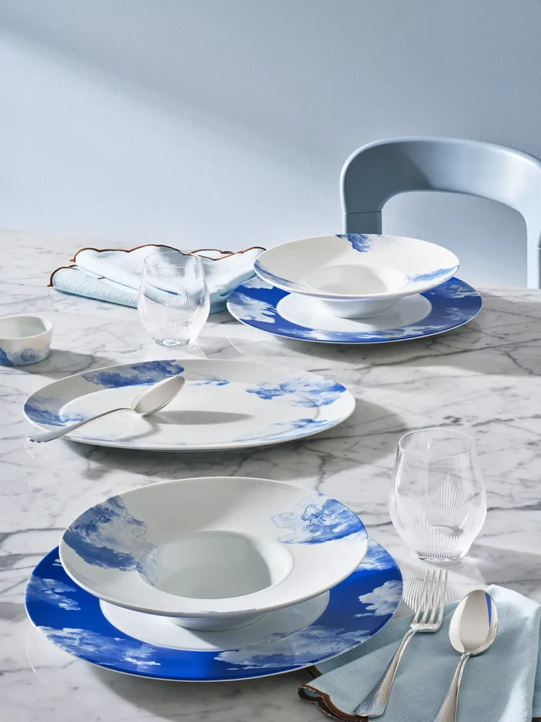 Assiettes à soupe en porcelaine RÊVERIE de Fürstenberg aux délicats motifs de nuages bleus, disposées sur une table en marbre avec des serviettes aux couleurs pastel, des couverts élégants et des verres galbés.