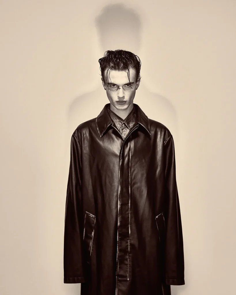 Stylized portrait of a male model in a dark leather coat with slicked hair and a dramatic shadow cast on the wall