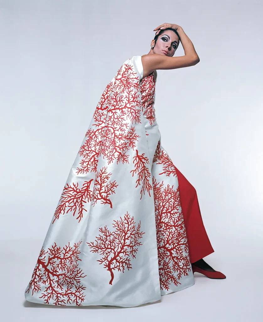 Studio portrait of a model wearing a white Valentino gown with red coral embroidery, paired with red trousers and bold eye makeup.