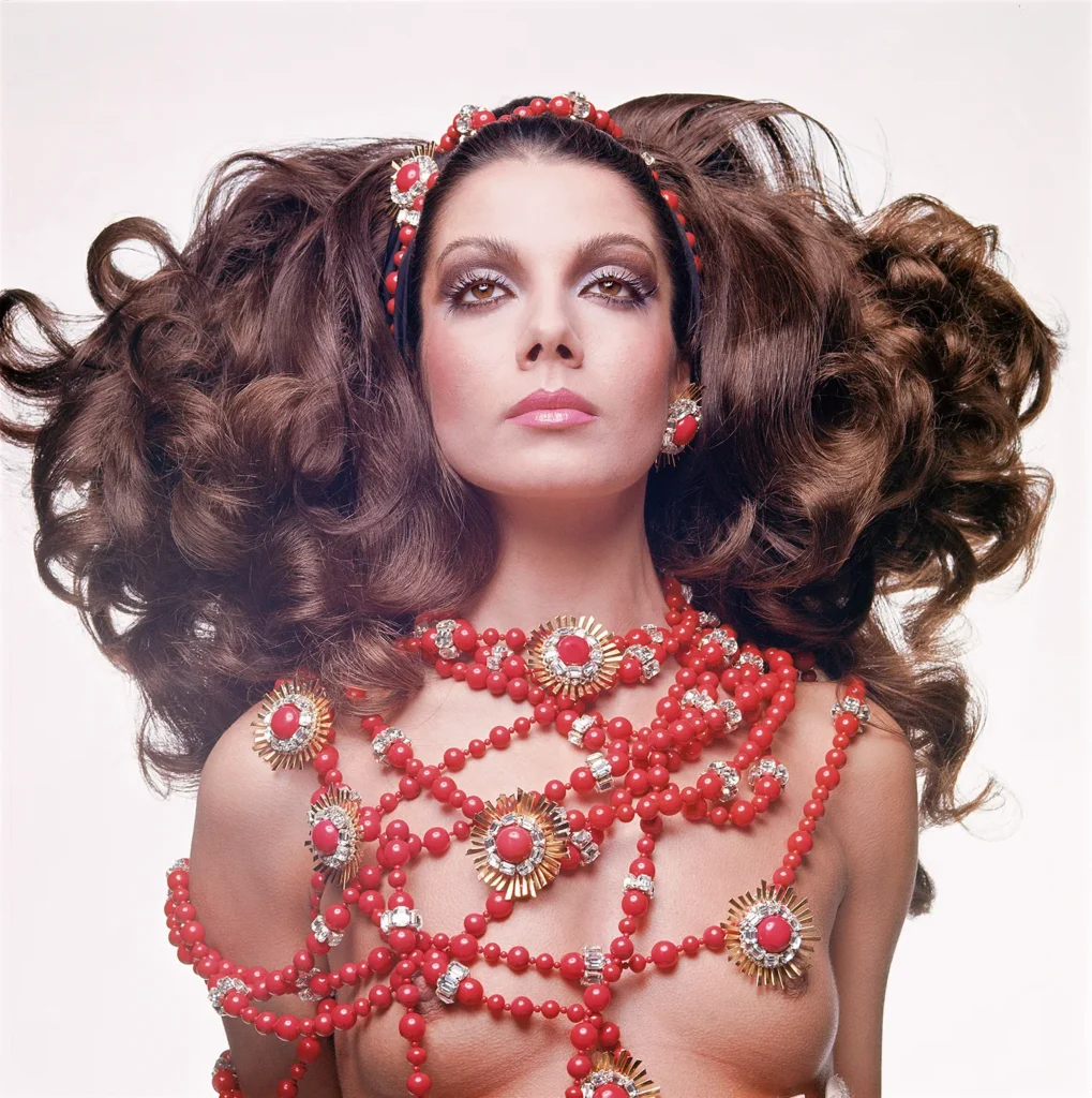Studio portrait of a model wearing elaborate red and gold Valentino jewelry and voluminous hair, photographed against a white background.