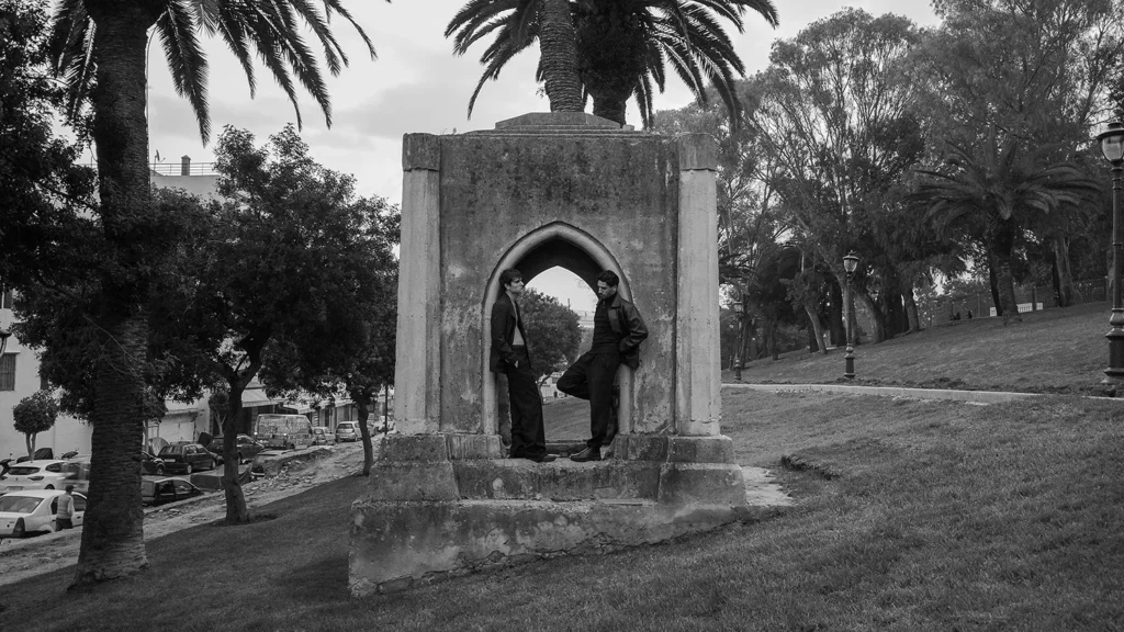 Two men in dark clothing framed by a stone arch in a Tangier park, surrounded by palm trees and grass.